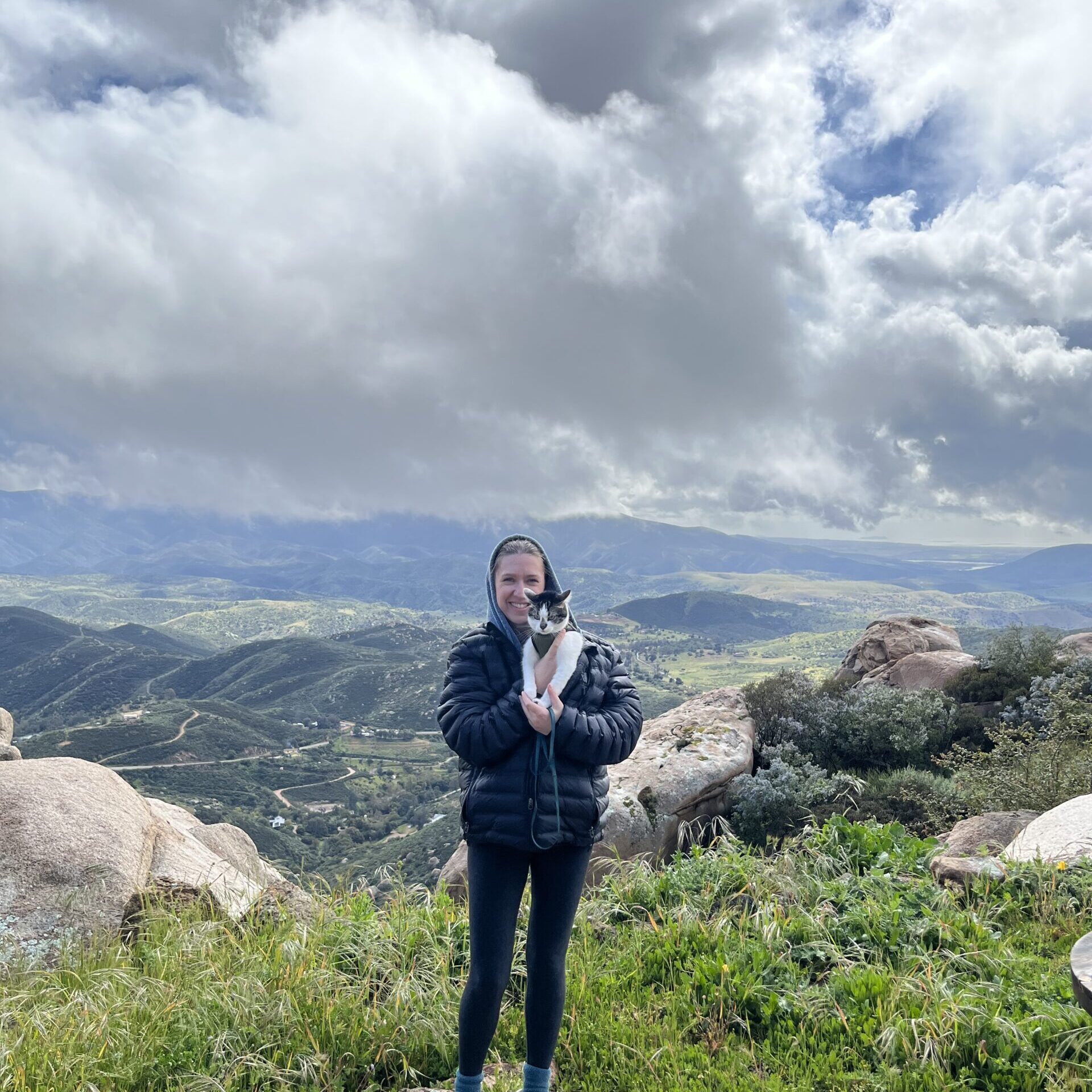 Photo of me in warm clothes, holding a white cat with black nose and ears, standing in front of a scenic background of hills and white fluffy clouds.